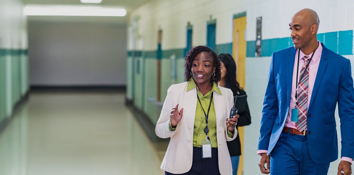 Two school administrators walking and talking in a brightly lit hallway. The woman in a white blazer and green shirt gestures while holding a walkie-talkie, and the man beside her wears a blue suit with a plaid tie.
